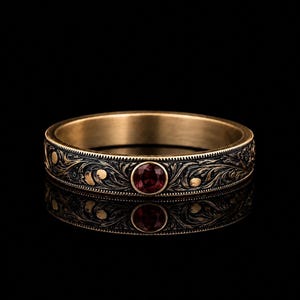 Könnte beinhalten: Ein goldener Bandring mit detaillierten, schwarz ausgefüllten Rankengravuren und einem einzelnen, rund gefassten, tiefroten Edelstein in der Mitte. Das Metall hat eine gebürstete Oberfläche mit polierten Kanten vor schwarzem Hintergrund.