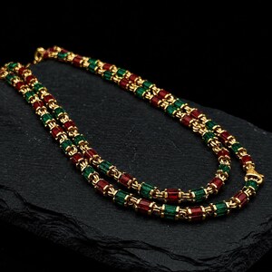 Può includere: Una collana a doppio filo con perline rettangolari rosse e verdi alternate, separate da piccoli distanziatori in metallo dorato. La collana presenta un motivo ripetitivo di queste perline colorate e accenti metallici lungo tutta la sua lunghezza.