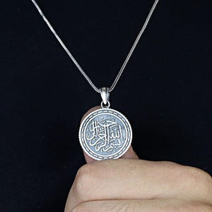 Op de afbeelding: Een zilverkleurige hangerketting met een cirkelvormige hanger met gegraveerd Arabisch schrift. De hanger hangt aan een fijne ketting. De ketting wordt getoond tegen een donkere achtergrond.