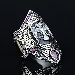 May include: A silver ring with an elaborate design, showcasing a central bird emblem with outstretched wings. The ring is adorned with small, round, and teardrop-shaped red gemstones. The ring is set against a black background.