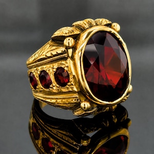 May include: A gold ring featuring a large, oval-cut, deep red garnet gemstone as the centerpiece. The band is crafted with intricate, textured metalwork and is accented by three smaller, round-cut red garnet stones along the side of the shank.