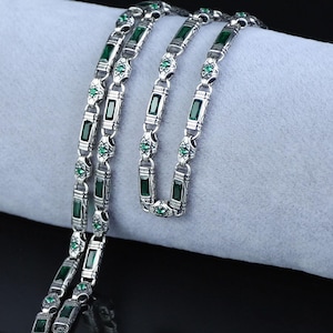 May include: A silver-toned necklace with rectangular and oval green gemstone accents. The necklace is draped over a soft, light gray surface, with a dark reflective background. The gemstones are emerald green, and the metal has an antique finish.