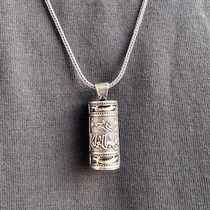 Könnte beinhalten: Eine silberne Anhängerkette mit einem zylindrischen Anhänger. Der Anhänger weist eingravierte Muster und Symbole auf und hängt an einer filigranen Kette. Die Halskette wird vor einem grauen Hintergrund präsentiert.