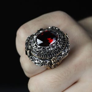 Peut inclure: Bague en argent ornée d'une grosse pierre rouge foncé de forme ovale. La bague présente des détails de filigrane complexes autour de la pierre et une bande avec des ornements supplémentaires. Un bijou de caractère.
