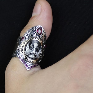 May include: A silver ring with an intricate design, featuring a central emblem and small red gemstones. The ring has an ornate, gothic style with gold accents and is worn on a thumb.
