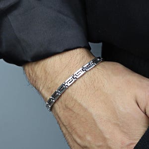 May include: A silver-toned bracelet with a geometric design is worn on a wrist. The bracelet is composed of rectangular links with a repeating pattern. The person is wearing a black long-sleeved shirt.