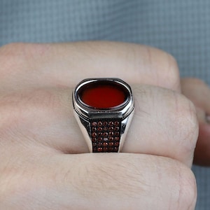 May include: A silver ring with a rectangular, deep red stone. The ring has a detailed design with small red stones around the base of the main stone. The ring is worn on a finger.