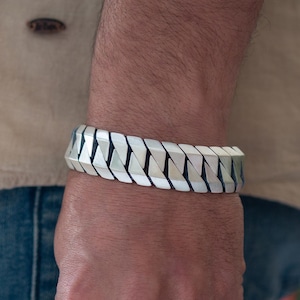 May include: A silver-toned bracelet with a geometric design is worn on a wrist. The bracelet is made of interlocking, angular pieces. The person is wearing a light-colored shirt and blue jeans, holding a jacket.