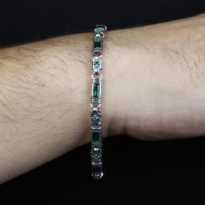 Op de afbeelding: Zilveren armband met rechthoekige en ovale smaragdgroene stenen. De armband is gemaakt van zilver en heeft een reeks afwisselende stenen. De armband wordt getoond op de arm van een persoon.