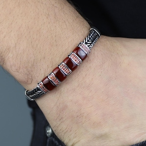 May include: Silver bracelet with a woven design and rectangular red stone accents. The bracelet features a series of polished red stones separated by smaller, patterned silver sections. The bracelet is worn on a wrist.