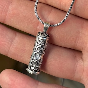 Peut inclure: Un collier pendentif argenté avec un pendentif cylindrique orné de motifs ajourés et d'un cœur. Le pendentif est suspendu à une fine chaîne en argent. Le design est détaillé et orné.
