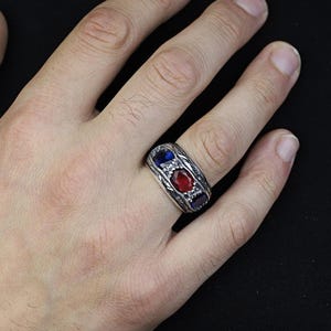 Könnte beinhalten: Silberfarbener Ring mit drei ovalen Edelsteinen: blau, rot und blau. Der Ring hat ein detailliertes, graviertes Design um die Edelsteine. Der Ring befindet sich auf einer Hand vor schwarzem Hintergrund.
