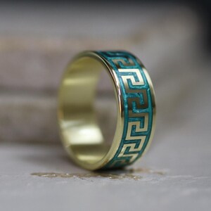 May include: A gold ring featuring a teal Greek key design around the band. The ring has a polished finish and a classic design. The teal colour provides a striking contrast against the gold.