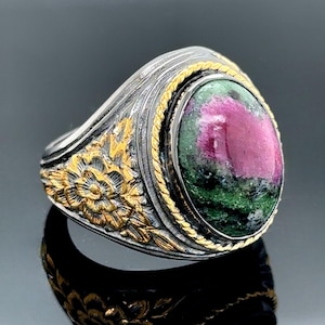 Puede incluir: Un anillo de plata con una piedra preciosa verde y rosa engastada en un bisel de oro. La banda del anillo está decorada con diseños florales.