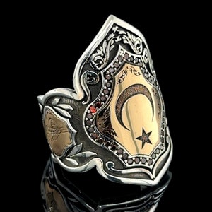 Może przedstawiać: Srebrny pierścionek z motywem złotego półksiężyca i gwiazdy. Pierścionek zdobiony jest czerwonymi kamieniami szlachetnymi i misternymi wzorami kwiatowymi.