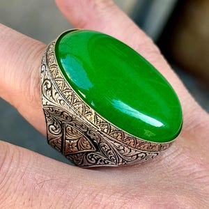 May include: A silver ring featuring an intricate design and a large, oval green gemstone.
