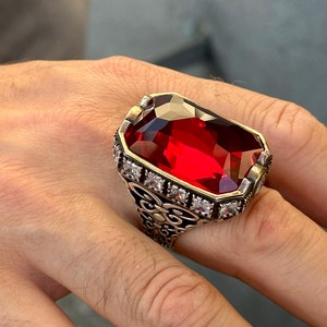 May include: A silver ring with a large, red, faceted gemstone surrounded by smaller, clear gemstones. The ring has an intricate, ornate design.