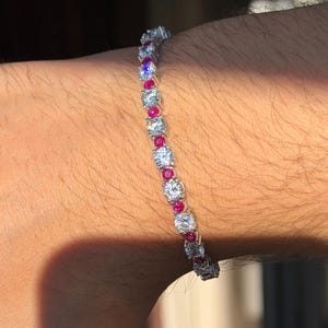 Op de afbeelding: Een tennisarmband met een herhalend patroon van afwisselend ronde heldere stenen en kleinere ronde rode stenen, gezet in een zilverkleurige metalen band. De armband heeft een flexibele schakelstructuur die om de pols sluit.