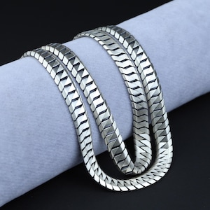 Op de afbeelding: Een zilverkleurige, platte slangenketting. De ketting is gemaakt van in elkaar grijpende geometrische segmenten, waardoor een textuurrijk, flexibel ontwerp ontstaat. De ketting wordt gepresenteerd op een lichtgrijze ondergrond tegen een donkere achtergrond.