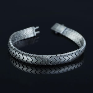 May include: A silver bracelet featuring a herringbone design. The bracelet is composed of interlocking, zig-zag shaped links. The clasp is open, showing the internal mechanism. The bracelet is presented on a reflective black surface.