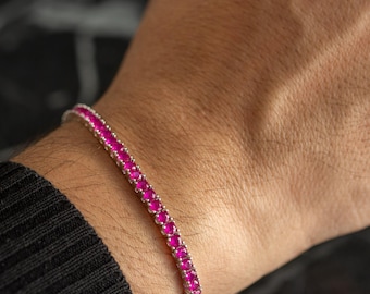 Peut inclure: Un bracelet de tennis argenté orné d'une rangée de pierres précieuses roses vives. Le bracelet est porté au poignet, mettant en valeur le design et la couleur du bijou sur un fond sombre.