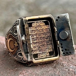 May include: A silver ring with a hinged compartment that opens to reveal a gold inscription of Arabic text. The ring also features a crescent moon and star design.