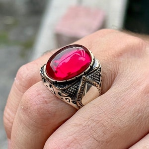 May include: A silver ring with an oval-shaped, bright red gemstone set in a detailed, ornate design.