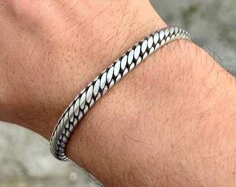 May include: A silver bracelet with a woven design. The bracelet is a solid band of metal with a repeating pattern of interlocking links. The bracelet is worn on a wrist.
