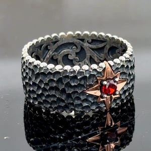 Może przedstawiać: Srebrny pierścionek z czarnym oksydowanym wykończeniem i czerwonym kamieniem szlachetnym osadzonym w gwiazdkowym wzorze z różowego złota.