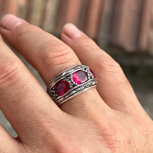 Op de afbeelding: Een zilveren ring met drie rode edelstenen ingelegd in een gedetailleerd bloemmotief.