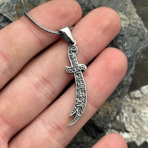 Può includere: Una collana d'argento con un ciondolo a forma di spada d'argento dettagliata. La spada è curva e presenta disegni intricati sulla lama.