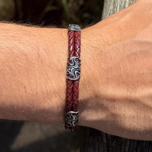 May include: A braided burgundy leather bracelet with silver-colored decorative accents. The bracelet features intricate metalwork designs and a secure clasp. This accessory is a stylish piece of jewelry.