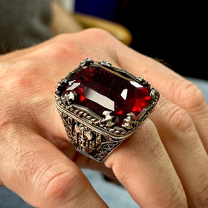 Op de afbeelding: Een zilveren ring met een grote, donkerrode edelsteen gezet in een gedetailleerd, versierd ontwerp.