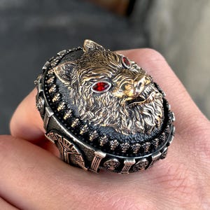 May include: A detailed silver ring featuring a boar's head with red gemstone eyes. The boar's head is gold-toned, set against a black background, and surrounded by an ornate silver band. The ring is a statement piece.