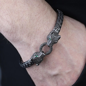 May include: A black panther bracelet with green eyes is shown. The bracelet is made of a dark metal and features two panther heads that connect to form a clasp. The bracelet is worn on a wrist.