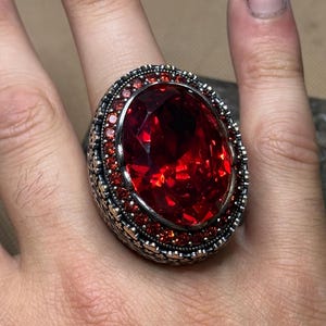 Peut inclure: Une bague ornée d'une grande pierre rouge facettée. L'anneau argenté est orné de petits brillants rouges et de motifs complexes. Un bijou audacieux, idéal pour les occasions spéciales.
