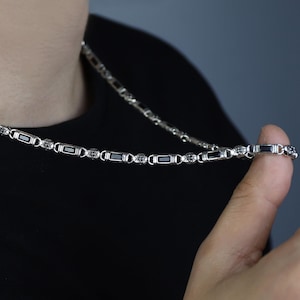 May include: A silver chain necklace with rectangular and circular links. The necklace is worn around the neck and held by a hand. The chain has dark accents within the links.