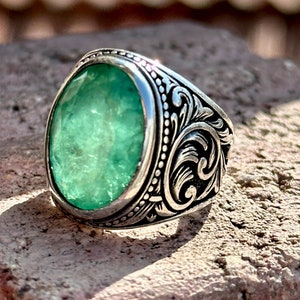 May include: A silver ring with an ornate design and a large, oval-shaped green gemstone.