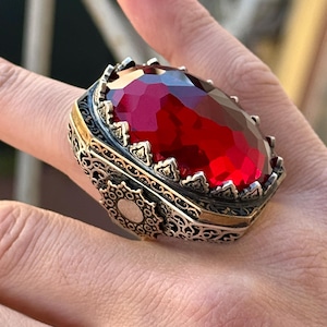 May include: A silver ring featuring intricate filigree detailing and a large, oval-shaped red gemstone set in a crown-like setting.