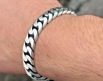 May include: A silver-colored curb chain bracelet. The bracelet is made of interlocking links, creating a bold and textured appearance. The bracelet is worn on a wrist, showcasing its design and craftsmanship. The links are approximately 0.5 inches wide.