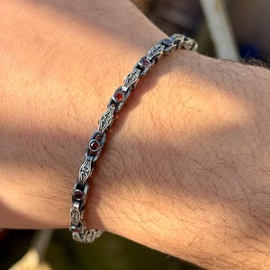 May include: A silver bracelet with a detailed, ornate design. The bracelet features small, round, red gemstones set within the links. The bracelet is worn on a person's wrist, showcasing its intricate craftsmanship and elegant style.