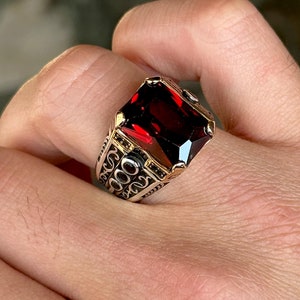 Può includere: Un anello d'argento con una grande pietra preziosa rossa di forma quadrata. L'anello è decorato con disegni intricati e dettagliati attorno alla pietra preziosa.