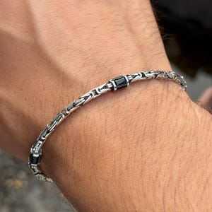 May include: A silver bracelet featuring a detailed chain design with black rectangular accents. The bracelet is displayed on a wrist, highlighting its intricate links and dark gemstone-like features. The image provides a close-up view of the jewellery.