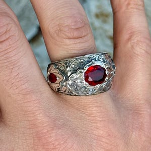Può includere: Anello in argento ornato con una grande gemma rossa ovale al centro e pietre rosse più piccole sui lati. L'anello presenta dettagliate incisioni floreali e a volute, che mostrano uno stile vintage o antico. L'anello è mostrato su un dito.