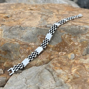 Könnte beinhalten: Ein silbernes Armband mit einem Schachbrettmuster in Schwarz und Weiß. Das Armband besteht aus Metallgliedern und hat einen Verschluss.