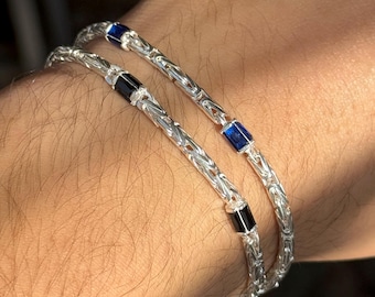 Könnte beinhalten: Zwei silberne Armbänder mit einem gewebten Kettendesign. Jedes Armband hat rechteckige blaue und schwarze Edelsteinakzente. Die Armbänder werden an einem Arm gezeigt und zeigen die Details des Schmucks.