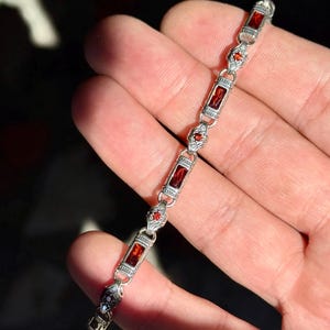 Può includere: Un braccialetto d'argento con pietre preziose rosse rettangolari e a forma di diamante. Il braccialetto è tenuto in una mano, con la luce del sole che si riflette sul metallo e sulle pietre. È un gioiello.