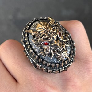 Puede incluir: Un anillo de plata ornamentado con un diseño detallado de cara de demonio en color dorado. El anillo presenta ojos de piedras preciosas rojas y patrones intrincados, sobre una base ovalada. El anillo tiene un acabado oscuro oxidado.