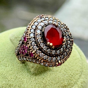 May include: A silver ring with a large red gemstone surrounded by smaller white gemstones. The ring has a detailed design with intricate filigree work.