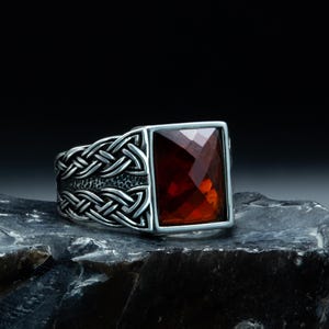 May include: A silver ring with a rectangular, faceted, red gemstone. The ring features a Celtic knot design on the band. The ring is displayed on a dark, textured surface against a black background.